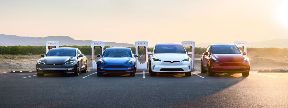 Tesla Model S, Model 3, Model X and Model Y lined up at Supercharger posts at sunset