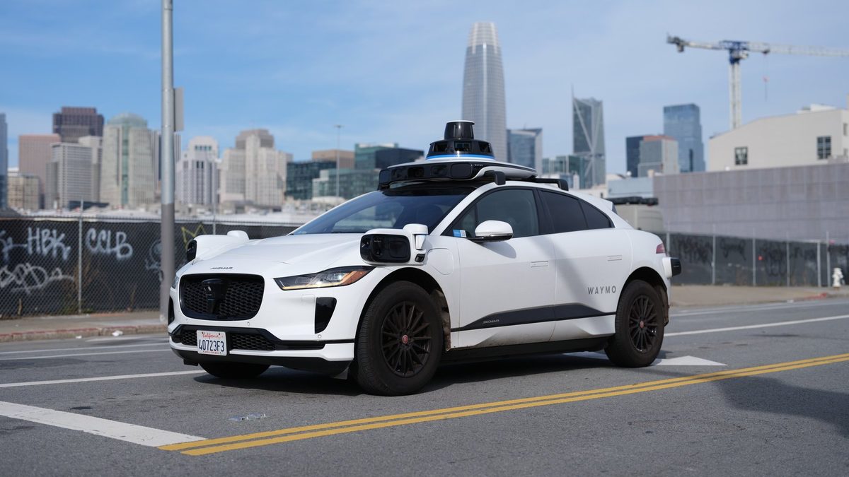 White Waymo Jaguar I-Pace with nobody at the wheel on a San Francisco street, with the city's skyscrapers in the background